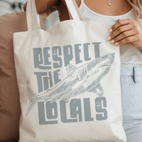 Woman holding Respect the Locals Shark Canvas Tote Bag supporting shark conservation, 100% organic cotton, 15x16in size, twin carry handles.