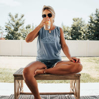 Woman in Stay Salty Surfboard Blue Jean Tank sitting outdoors with a drink